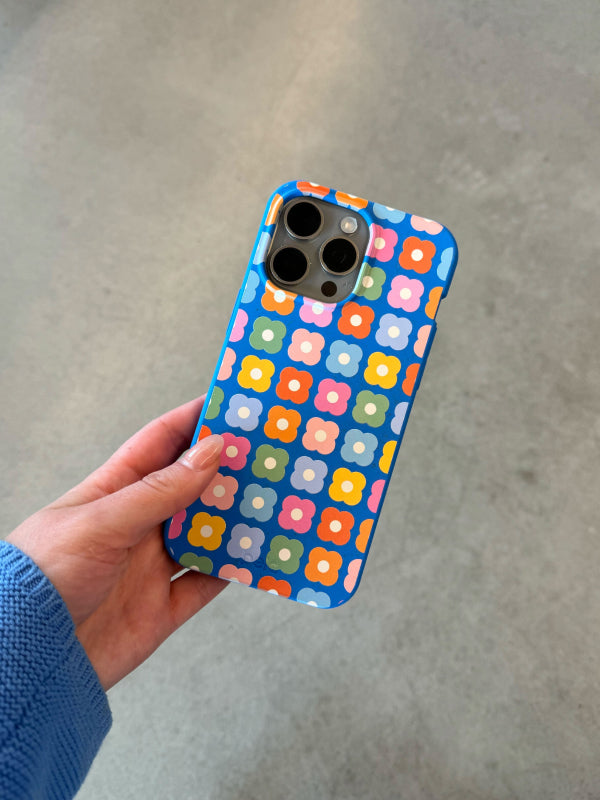 A hand holding the Pela Petal Pop Phone Case from the 60s Collection. The case features a vibrant pattern of colorful flowers in pink, orange, yellow, green, and blue against a blue background. The person's nails are painted light pink, and they are wearing a blue sweater. The background is a neutral, light-colored surface.