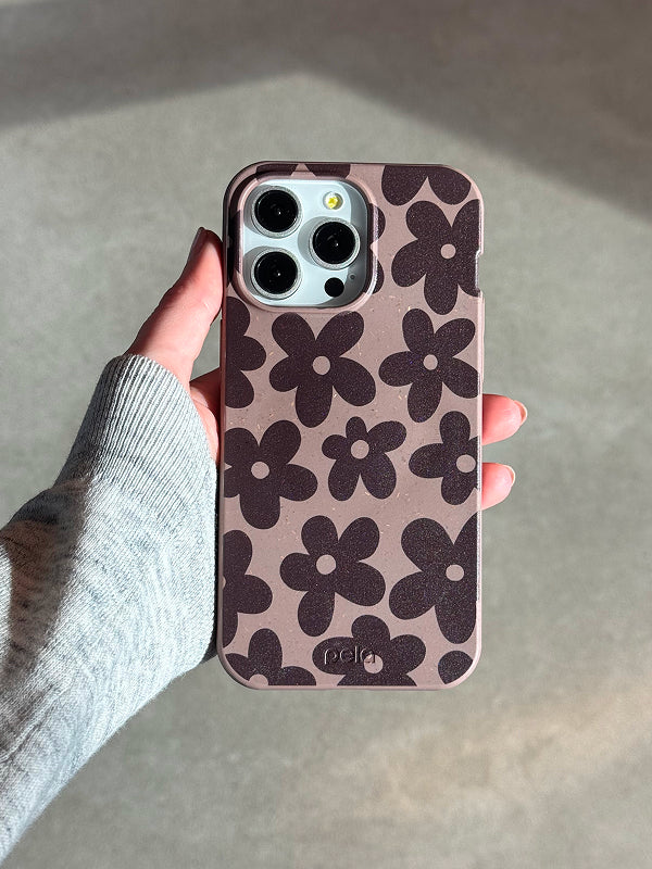 A hand holds a Pela Earthy Blooms phone case in a warm, earthy brown shade with a dark brown floral pattern featuring bold, retro-inspired flowers. The case has a smooth, matte texture with visible speckles, highlighting its eco-friendly, compostable material. The phone inside has a triple-lens camera, and the person holding it is wearing a light gray sweater. The natural lighting casts soft shadows, emphasizing the case’s stylish and sustainable design.