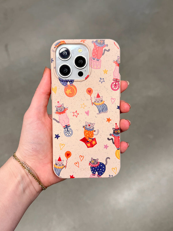A hand holds a Pela phone case with a beige, speckled background featuring a playful circus-themed design of illustrated cats dressed as clowns. The cats are depicted juggling, riding unicycles, popping out of jack-in-the-boxes, and holding balloons, surrounded by colorful stars and hearts. The case has a soft, matte texture and is eco-friendly, made from compostable materials. The phone inside has a triple-lens camera.
