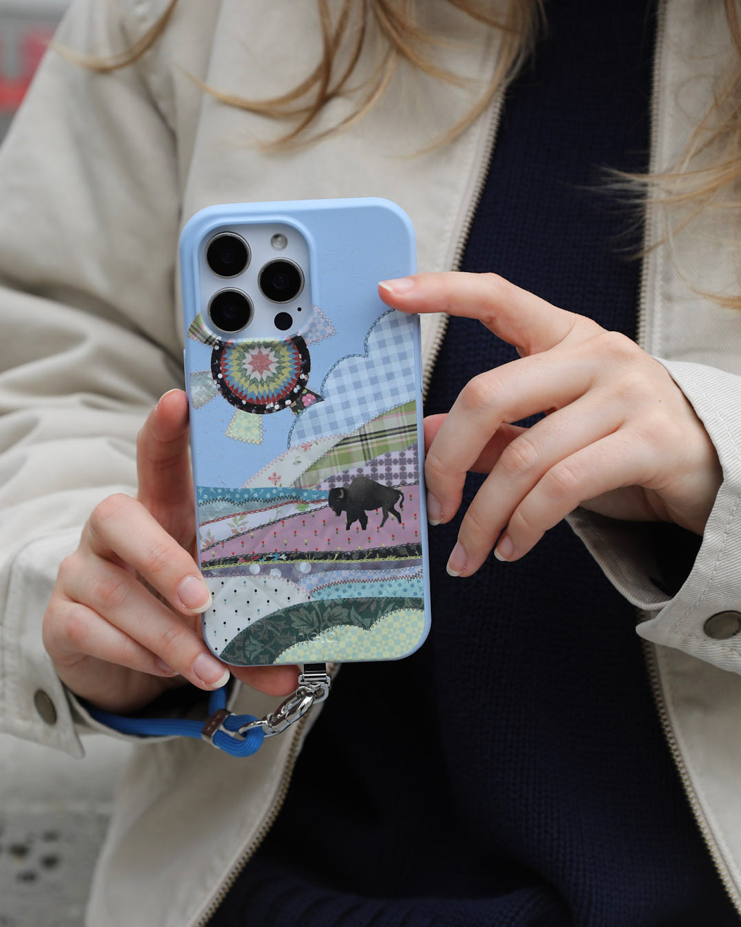 A woman holding a Pela Quilted Prairie phone case in Powder Blue featuring a patchwork quilt landscape scene with a silhouetted bison, gingham clouds, a crochet medallion hot air balloon, and layered fabric-print rolling hills in soft pastels and florals, with a blue Pela phone strap attached — wearing a beige trench coat over a dark navy knit sweater.