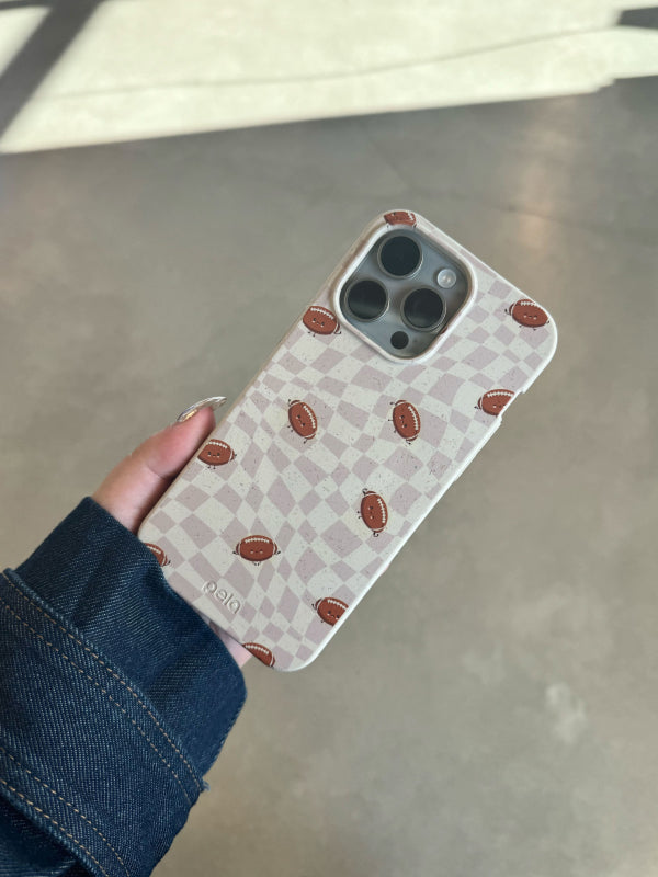 A hand holds the Pela Football Groove Phone Case, which features a light beige and white checkered pattern with small brown footballs scattered across the design. The eco-friendly case is sleek and stylish, perfect for sports enthusiasts. The person’s denim jacket contrasts with the case, creating a casual, sporty vibe.