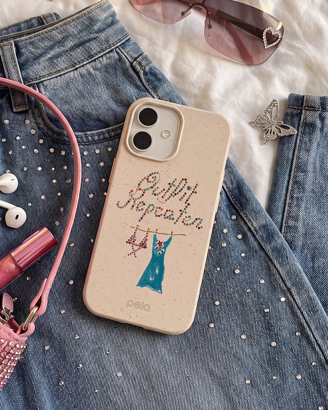 A Pela Outfit Repeater phone case in Seashell featuring rhinestone-style lettering and an illustrated blue dress on a hanger, styled on a rhinestone-studded denim jacket alongside a pink phone strap, white earbuds, pink lip gloss, rimless tinted sunglasses, and a crystal butterfly brooch.