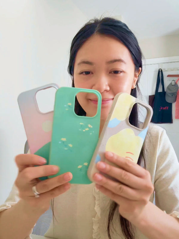 Katie Waiyu holding up three phone cases she designed for the Pela Artist Series Collection, featuring her signature pastel landscapes and whimsical style.