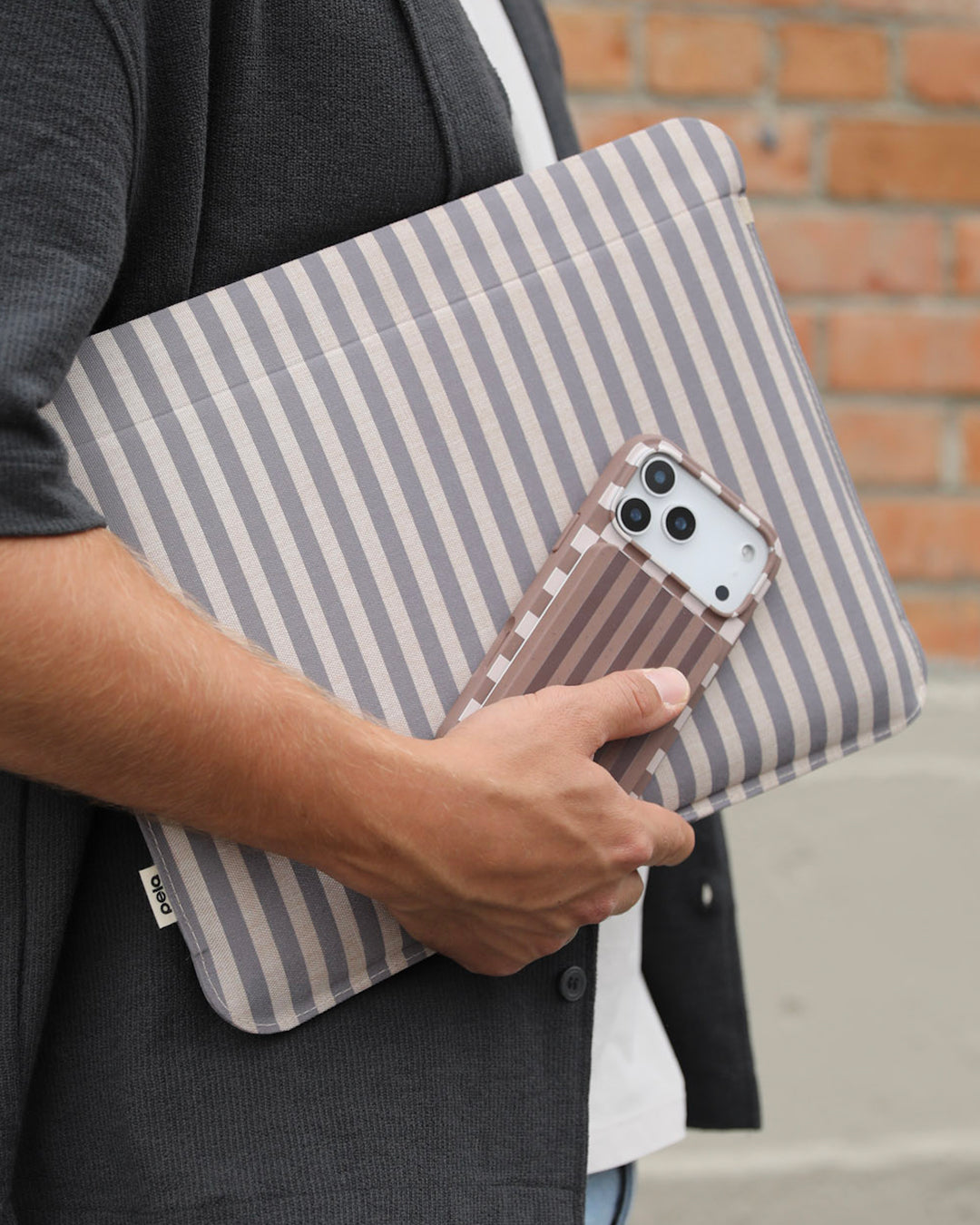 A man carrying a Pela Fawn Stripe Laptop Sleeve in taupe and cream vertical stripes tucked under his arm, holding a matching Pela phone case in the same tonal colourway — wearing a dark grey cardigan over a white t-shirt with a brick wall in the background.