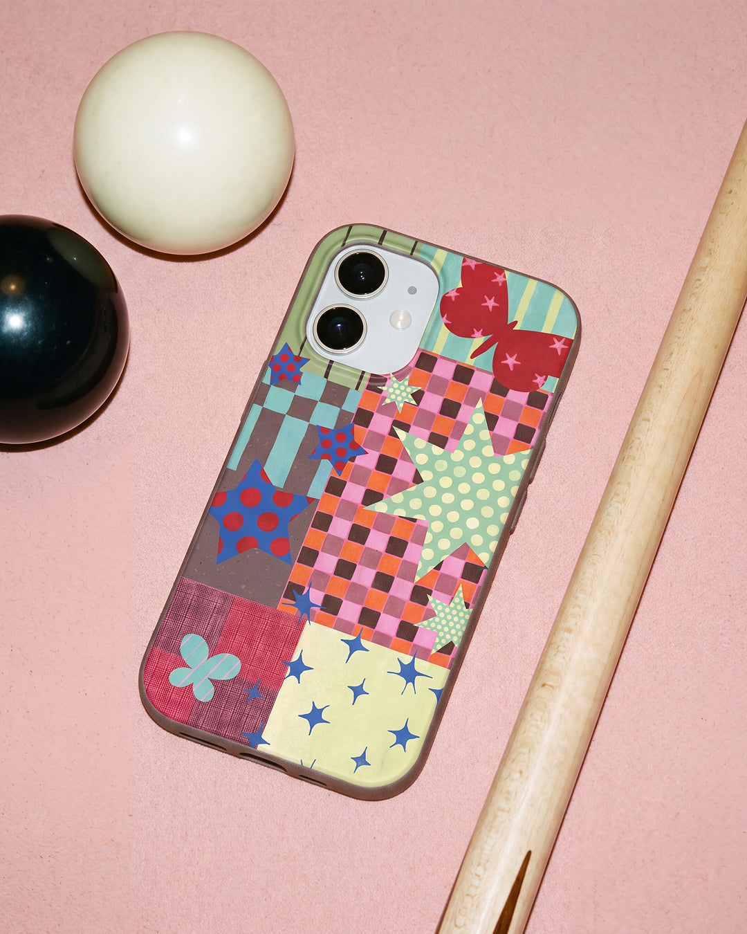 A Pela Patchwork Journal phone case featuring a colourful patchwork collage of plaid, gingham, and checker patterns with stars, butterflies, and floral motifs in pink, blue, green, and yellow, resting on a pink pool table surface alongside a white cue ball, black billiard ball, and wooden pool cue.