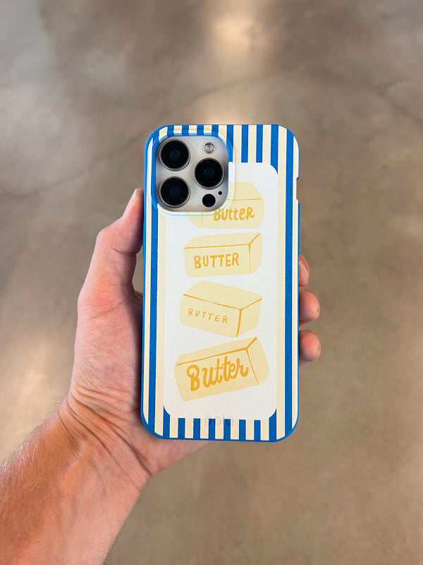 Hand holding the Butter Bars Phone Case by Pela, featuring playful yellow butter stick illustrations stacked on a white background with bold blue stripes.