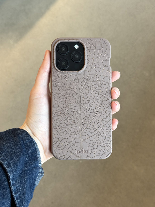 A hand holds a Pela phone case in a muted, earthy brown color featuring an intricate embossed leaf vein pattern. The detailed texture mimics the natural structure of a leaf, creating an organic and tactile design. The case is made from Pela’s signature compostable material, offering a sustainable and stylish choice. The phone inside has a triple-lens camera, and the person holding it is wearing a dark denim jacket. Soft natural lighting highlights the case’s unique texture and eco-friendly appeal.