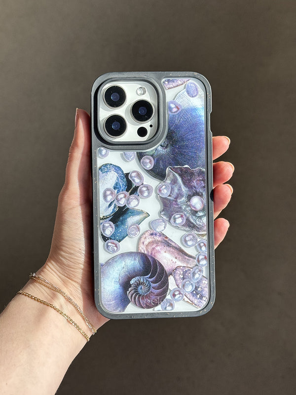 A hand holds a Pela phone case featuring the Deep Sea Dreams design. The clear case allows the phone’s color to show through, overlaid with iridescent seashells, nautilus shapes, and scattered pearls in soft shades of purple, blue, and pink. The design creates a dreamy, ocean-inspired effect while maintaining visibility of the device. The phone inside has a triple-lens camera, and the case is outlined with a translucent gray border.