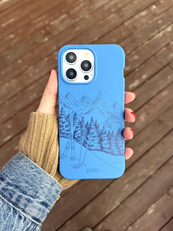 A hand holds an iPhone featuring the Pela Backcountry Ski phone case, which showcases a detailed blue design of a mountain landscape with skis and snow-covered trees. The background is a natural wooden deck, adding warmth to the scene. The person is wearing a denim jacket and a tan knit sweater, with red nail polish that complements the wintery, earth-friendly aesthetic of the phone case.