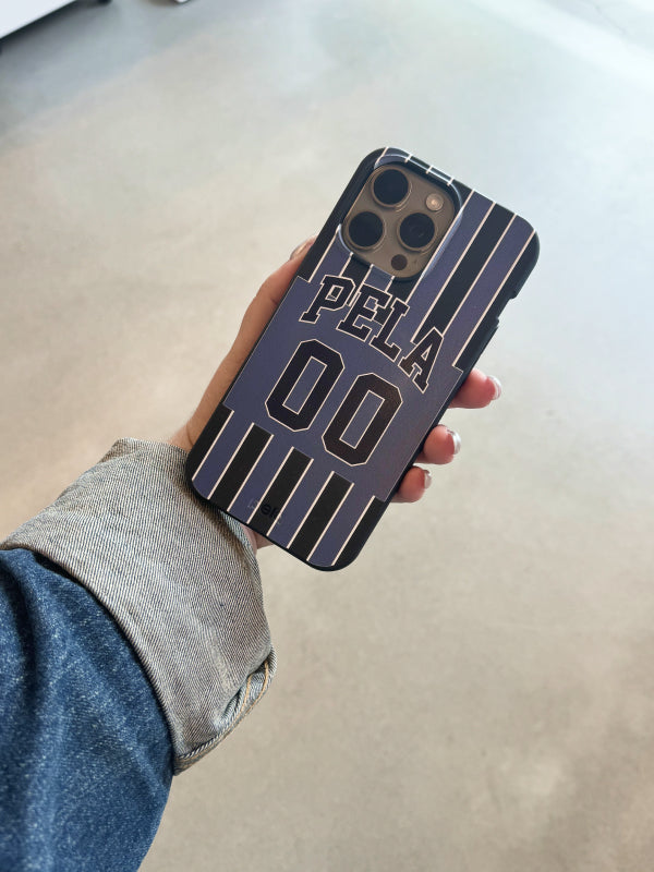 A hand holds the Pela Soccer Jersey Phone Case, featuring a design reminiscent of a classic soccer jersey. The case is adorned with vertical stripes in dark blue and white, with the text “PELA” and the number “00” prominently displayed in the center, mimicking a player’s jersey number. The overall look exudes a sporty and stylish vibe, perfect for soccer enthusiasts.