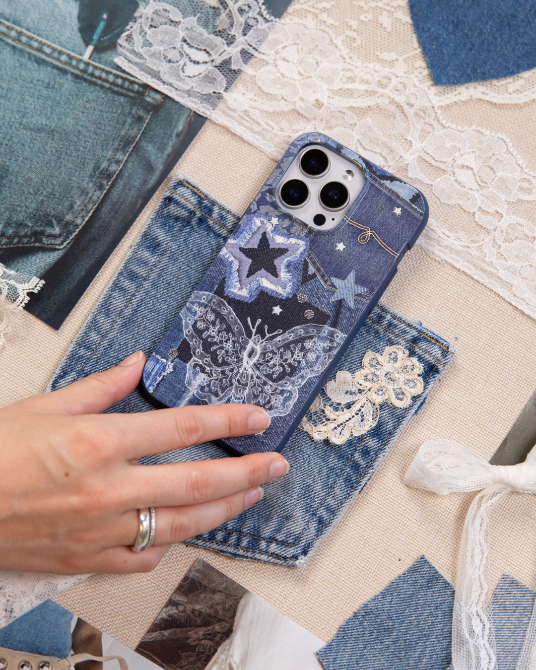 A hand holding a Pela Double Denim phone case featuring a patchwork denim collage design with an embroidered-style lace butterfly, denim stars, and scattered sparkles in shades of blue, resting on a flat lay of denim fabric swatches, lace trim, and ribbon on a natural linen background.