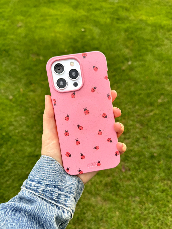 A hand is holding an iPhone featuring the Pela Rosy Ladybug phone case against a backdrop of bright green grass. The case has a playful design of small red ladybugs scattered across a soft pink background. The vibrant, earth-friendly case is paired with a casual denim jacket sleeve, highlighting its fun and nature-inspired theme.