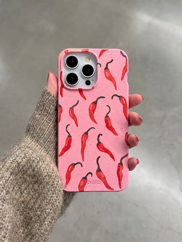  hand holding the Pela Heatwave Phone Case, featuring a vibrant pink background adorned with red chili peppers. The eco-friendly case, made from compostable materials, adds a bold and spicy touch to any phone. The person is wearing a cozy beige sweater, with a neutral-toned background enhancing the case's eye-catching design.