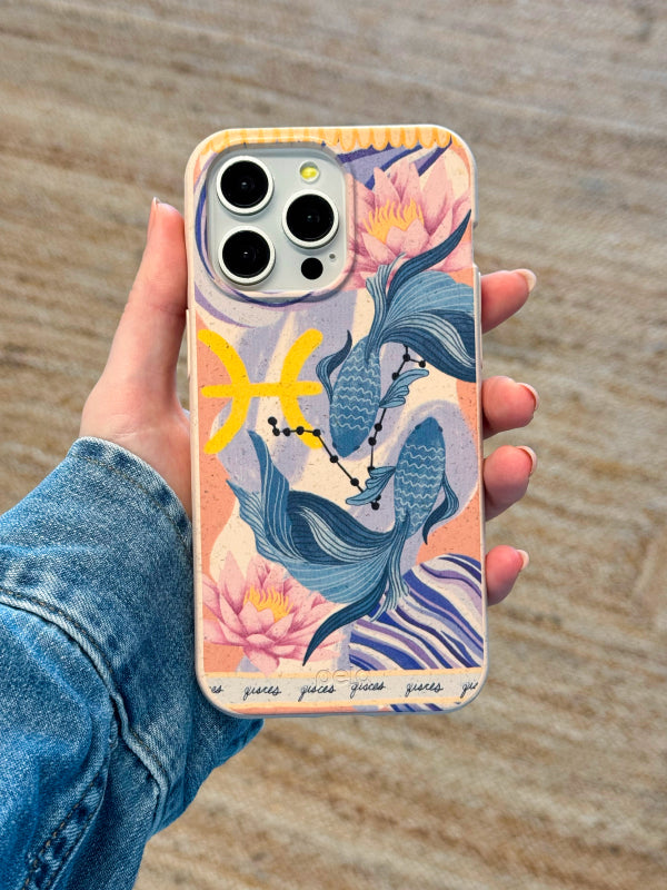 A hand holds the Pela Pisces Fish phone case, featuring a dreamy design of two flowing fish encircled by lotus flowers, celestial details, and the Pisces zodiac symbol. The artistic blend of pastel blues, yellows, and pinks creates a tranquil, water-inspired aesthetic, perfect for astrology lovers. Set against a neutral indoor backdrop, the image highlights the case's vibrant design and earth-friendly appeal.