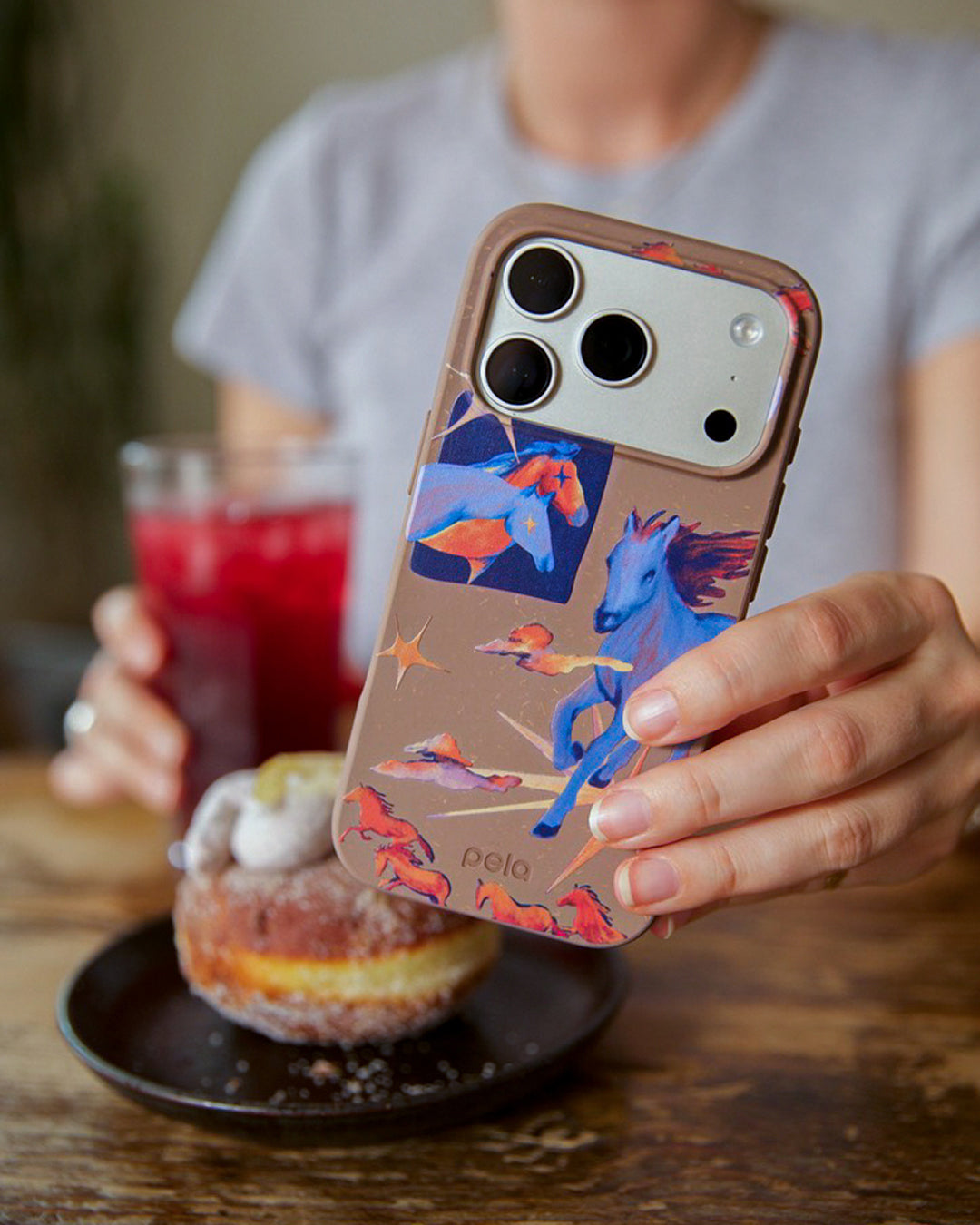 A woman holding a Pela Horse Dreamers phone case in taupe featuring illustrated blue and orange wild horses in motion with gold stars and dramatic clouds, held up at a café table beside a sugar-dusted doughnut on a black plate and a glass of red juice in the background.